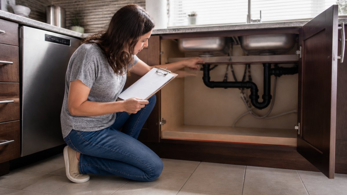 Homeowner standing in a kitchen with clipboard in hand, reviewing notes while pointing to under-sink pipes and fixtures, preparing the space for a service visit